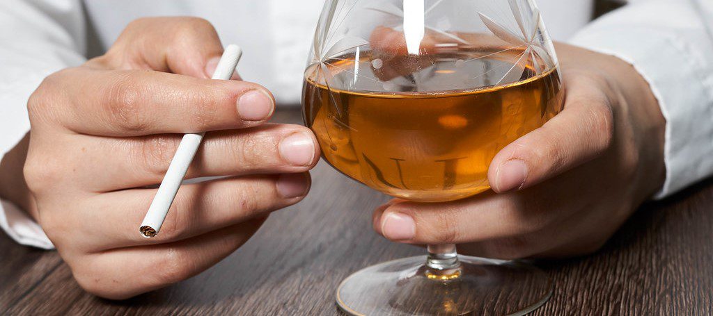 a person's hands, one holding a cigarette and the other holding a glass of what appears to be whiskey or another amber-colored spirit.