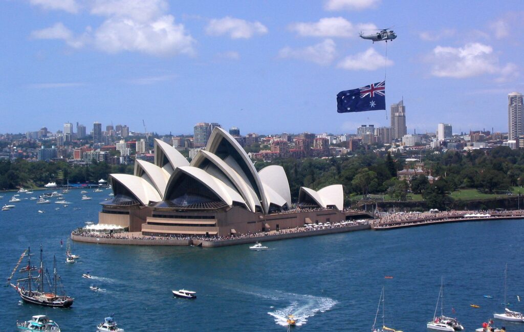 A celebratory scene, featuring the iconic Sydney Opera House with a large Australian flag being flown overhead by a helicopter.